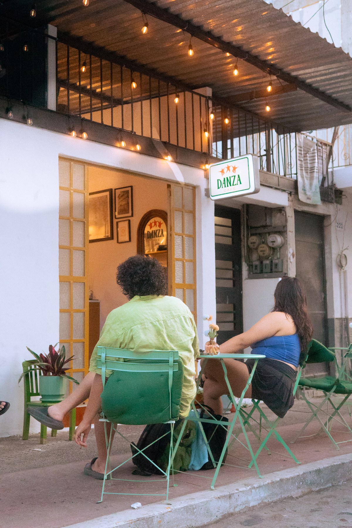 Atmospheric outdoor twilight patio at Danza Café in Puerto Vallarta with patrons seated at green bistro tables under warm string lights and illuminated lightbox blade sign
